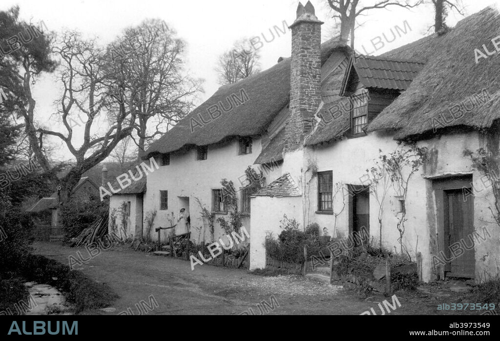 A pair of cottages with thatched roofs at Luccombe, Somerset, c1900. The cottage to the right has a dormer window and front chimney stack. The road outside is unpaved.