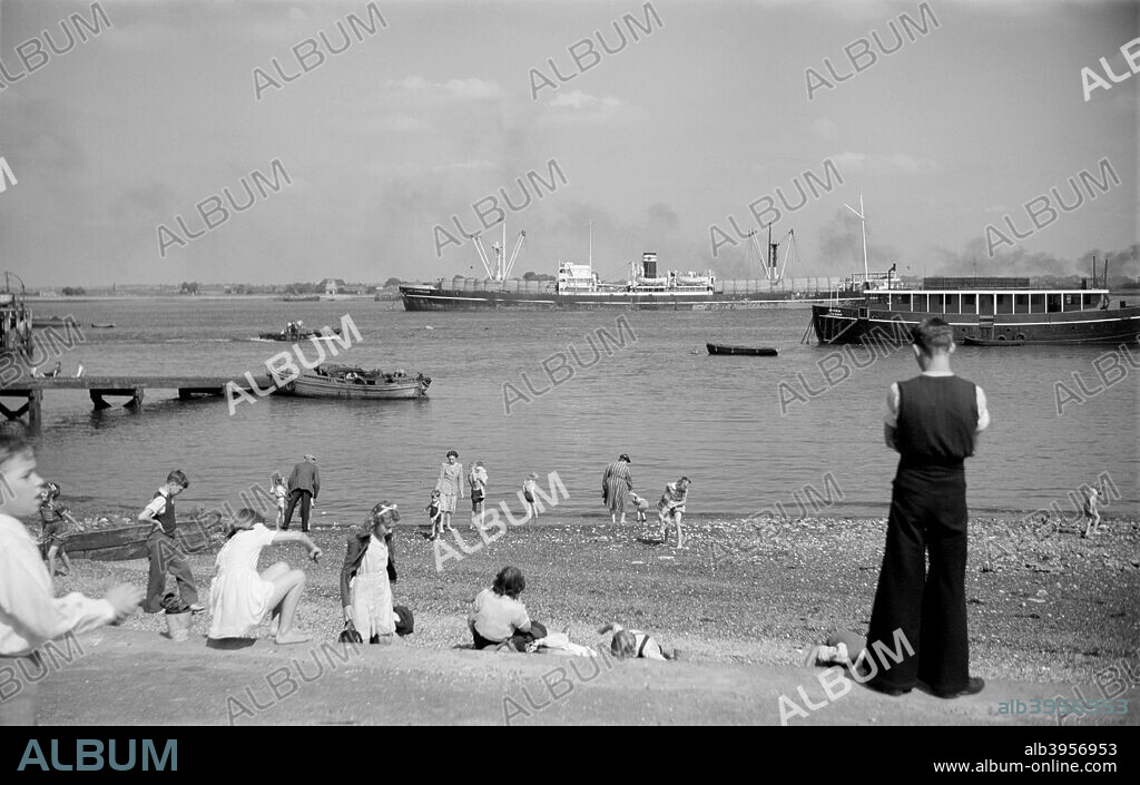 People on the beach at Gravesend, Kent, c1945-c1965. Children play on the beach at Gravesend while cargo vessels sail close in-shore. A boy in very wide flared trousers stands watching.