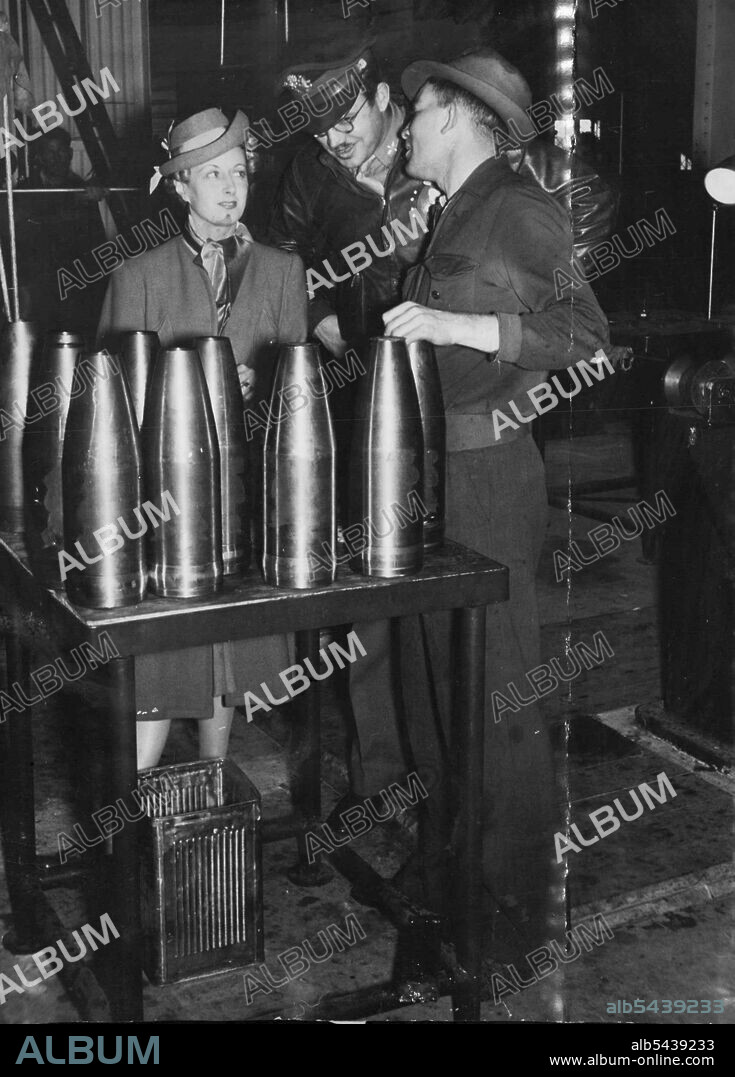 She'll Design Overalls -- Miss Mavis Ripper, dress designer, who will design overalls for the women who will shortly be taken on at Stewarts and Lloyds Newcastle factory, inspecting some of their shell cases with the koala stencil attached. The same koala design will be embroidered or stamped on the pocket of the girl's overalls. Talking to Miss Ripper are machine operator J. Uriah, of Hamilton, Newcastle, and U.S. airman Lieut. Holst of Pittsburgh. September 14, 1942.;She'll Design Overalls --.