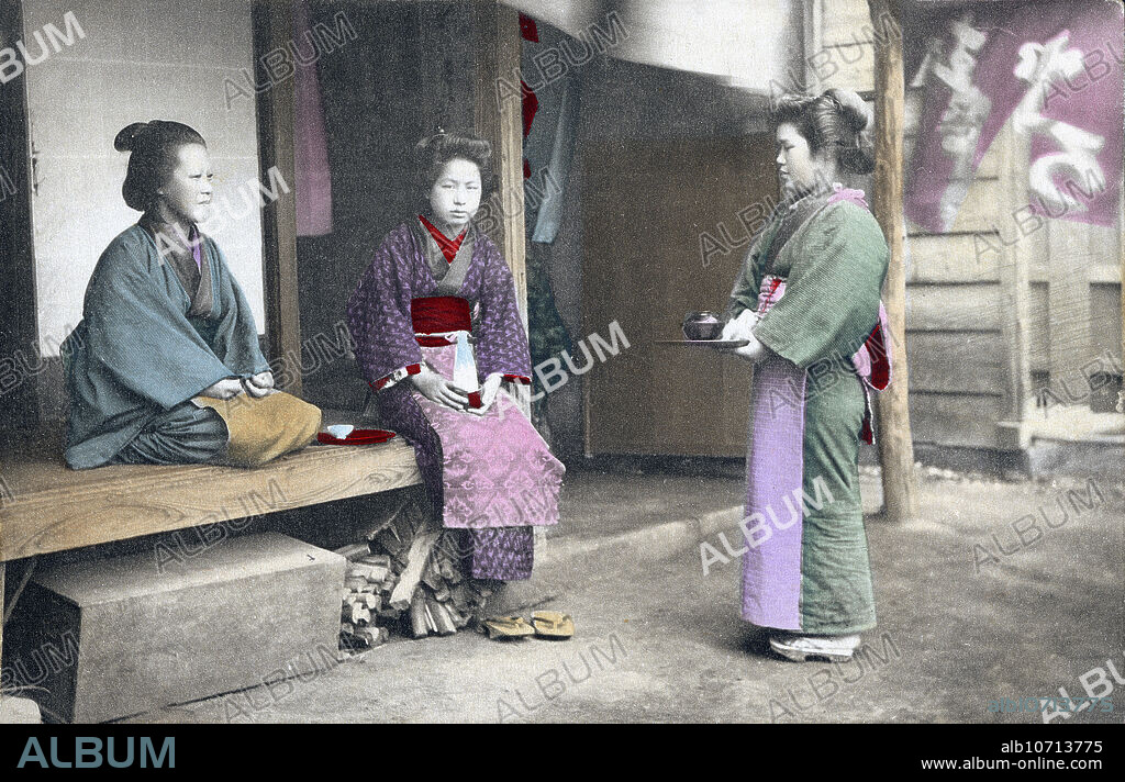 [ 1910s Japan - Japanese Maid Servant ] A maid servant in kimono, apron and traditional hairstyle at a tea house. She is holding a tray with a bowl. Two women sit on the engawa (porch). 20th century vintage postcard.