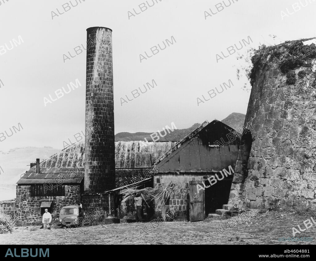 Monserrat, A sugar mill in MontserratA brick chimney and outbuildings form part of a sugar mill that was once the island’s principal rum factory. Two men can be seen carrying harvested sugar cane into one of the buildings. Montserrat, 1965, 1965. 2005/010/1/13/67.