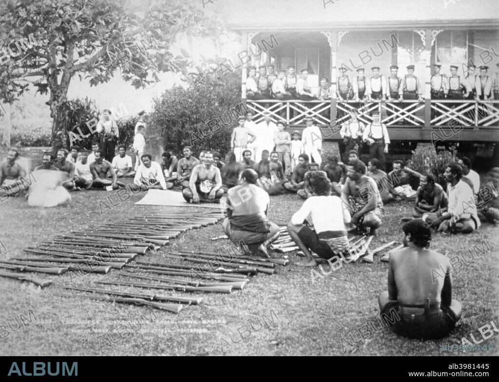 Tamasese distributing arms, Apia, Samoa, 1899. Samoan Civil War 1898-1899. A view showing Tamasese sitting on the ground, with rifles in front of him, and people sitting around. British sailors stand on the verandah of the building in the background.