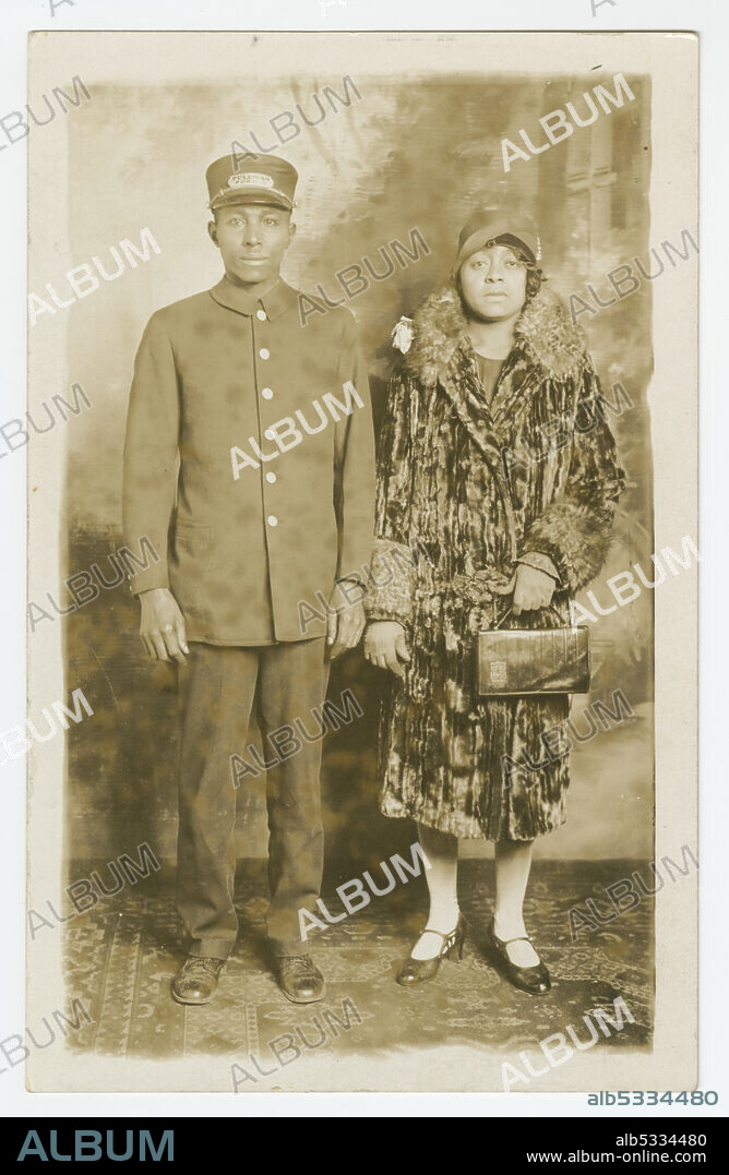 Black-and-white photographic postcard that was never stamped and mailed but has written in pencil on the back the name "Omer Ester". The image on the postcard is of Pullman Porter, Omer Ester, in his full porter uniform, including Pullman Porter cap. Standing next to him is an unidentified African American woman wearing what appears to be a velvet full length coat with fur trim on the collar and sleeves. She is wearing a 1920s style woman's hat and holding a handbag in her right hand. They are in a photography studio standing in front of a painted backdrop with an oriental-style carpet under their feet.
