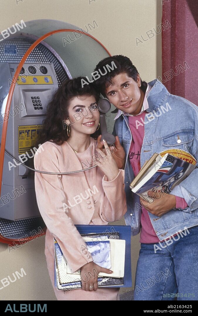 Cristina D'Avena and Marco Bellavia at a public phone. Italian actors Cristina D'Avena and Marco Bellavia are standing close by a public phone, lending a ear to the handset; they're in the middle of the shooting of the serial Arriva Cristina, based on the fictionalized life of the Italian singer herself. Cologno Monzese (MI), Italy, 1988.