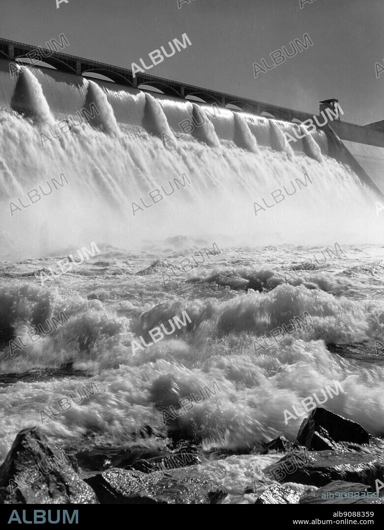 Grand Coulee Dam in Washington State, long one of the nations top tourist attractions, is the largest concrete structure ever built by man. The spray of twelve pipes in the centre of the picture are the outlets for the water pumped from the pool behind the dam into the Grand Coulee above. Here the water is stored for release in irrigating the million acre Columbia Basin Irrigation Project to the South. The dam is 46 stories high, higher than the Washington Monument. It is 12 city blocks long, 500 feet thick at its base. Now colour lighted at night, visitors are transfixed as 120 different colour combinations are played upon the dam during each half hour cycle brushing the massive drape of white water with their tints. The dam backs up the Columbia River 151 miles into Canada, to form Lake Roosevelt, a boaters paradise. The dam wa eight years building (1933 to 1941). The dam is the southern entrance to North Central Washington's Okanogan Highlands area. It is the first and largest of the series of great dams built or being built or planned for Columbia.