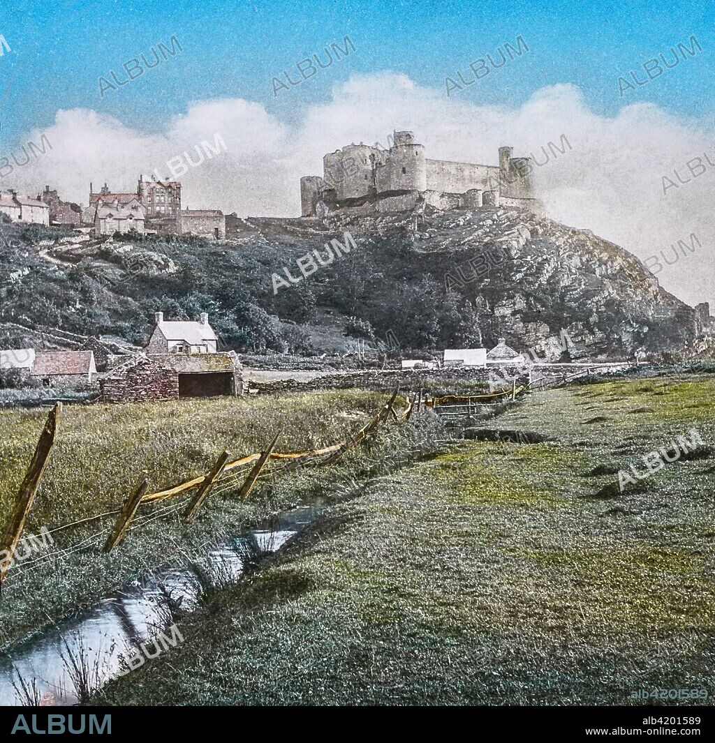 Magic Lantern slide circa 1900 hand coloured. created in 1887. A tour of North Wales. 39.Harlech Castle, from the Meadows.â€” This fine Castle is the remains of a once important fortress, situated on an elevated rock about half-a-mile from the sea. We are at once struck with the beauty of its situation and strength, a high perpendicular cliff, on the side nearest the sea, rendering it inaccessible. Like most of the other castles in the principality, this owes its erection to Edward I. Its siege bv Edward IV., when held by a Welsh warrior for the House of Lancaster, gave rise to that fine and popular air,  The March of the Men of Harlech.   A magnificent view of the Snowdonian range and CarÂ¬narvonshire mountains is obtained from the Castle itself, Harlech means bold rock. Ten miles further is.