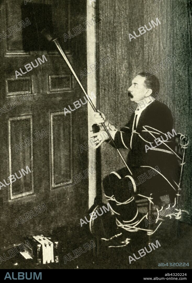 John Nevil Maskelyne performs a 'spirit cabinet illusion', c1910. 'How the tricks are performed - "A coach-horn or megaphone is useful for picking things up and putting them through the aperture in the door when securely tied'". John Nevil Maskelyne took up a challenge by Sir Hiram Maxim to reproduce a trick originally performed by the Davenport brothers, fraudulent spiritualists from America, and by an imitator of theirs, a Mr Fay. Maskelyne (1839-1917) was a British stage magician, escapologist, paranormal investigator and inventor of the pay toilet. From "Mr. Maskelyne's Reply to Sir Hiram Maxim", published in "The Strand Magazine, an illustrated monthly", Volume XL - July to December 1910. [George Newnes Ltd, London, 1910].