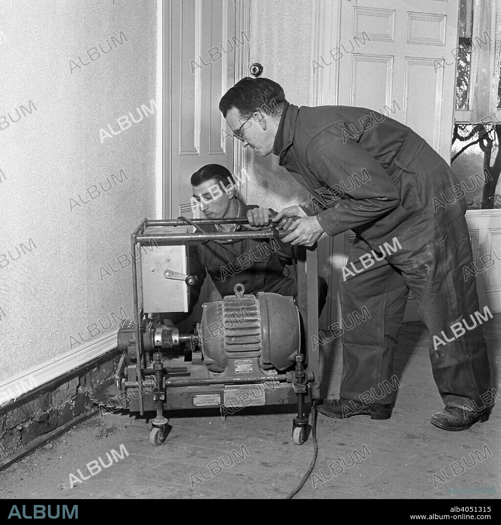 Damp proofing, Goldthorpe, South Yorkshire, 1957. Two workers use a cutting machine used to create a channel in which to insert a damp proof membrane. This photograph was taken for the Brooks Damp Proof Course Machine Company and the property being treated is in Furlong road in Goldthorpe. The 1950s and 1960s saw damp proofing installed in many houses which were originally built without any damp proof protection.
