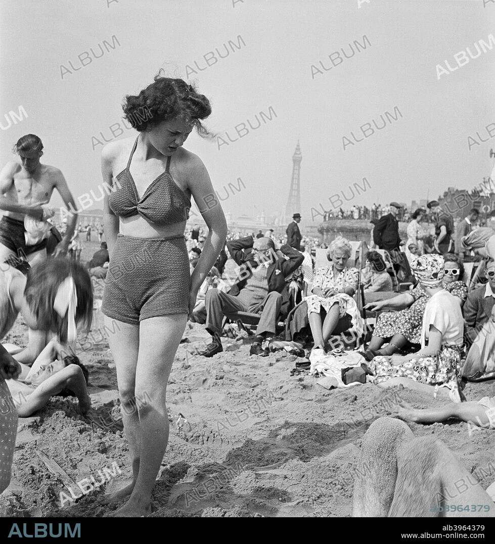 A young woman in a knitted bathing costume stands on the beach in the midst of the crowds of holidaymakers, Blackpool, c1946-c1955.  Blackpool Tower is visible in the background.