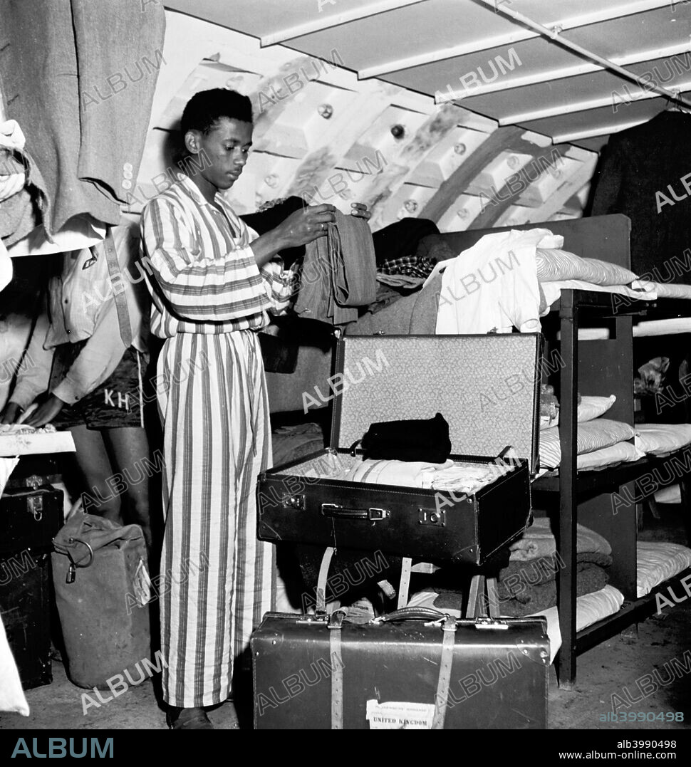 Immigrant living temporarily in a Nissen hut, London, 1950s. Inside a Nissen hut filled with bunk beds, a man dressed in pyjamas folds his clothes and puts them into a suitcase.