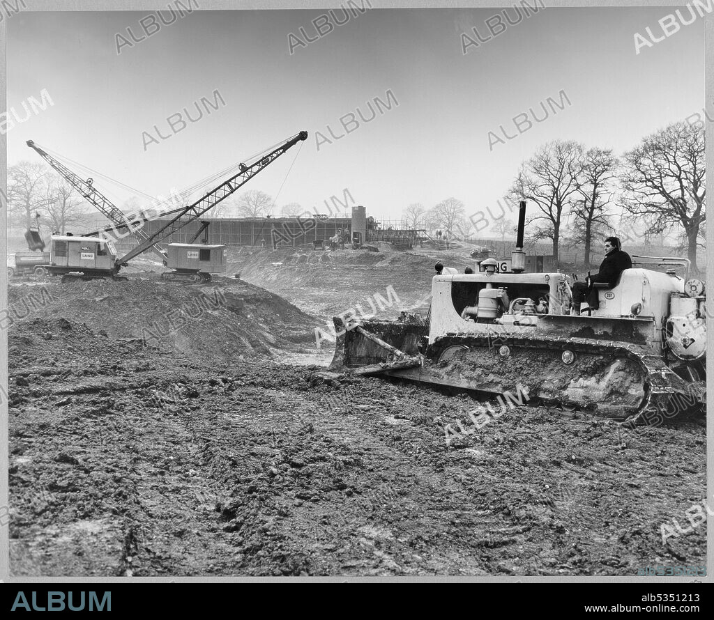 JOHN LAING PLC. A view of the construction of the Stafford By-pass on the Birmingham to Preston Motorway (M6), looking north across Rising Brook towards the Highfields Farm access bridge and showing Laing excavators and a bulldozer carrying out peat removal. In the album, the typed caption below the photograph reads 'Looking north across Rising Brook where large pocket of peat (50,000 cu.yds) is being removed and replaced with clay backfill prior to the motorway embankment being constructed. Background shows Highfields Farm access bridge, which is virtually complete.' The work on the Stafford By-pass started in June 1960 and was opened to traffic in August 1962.