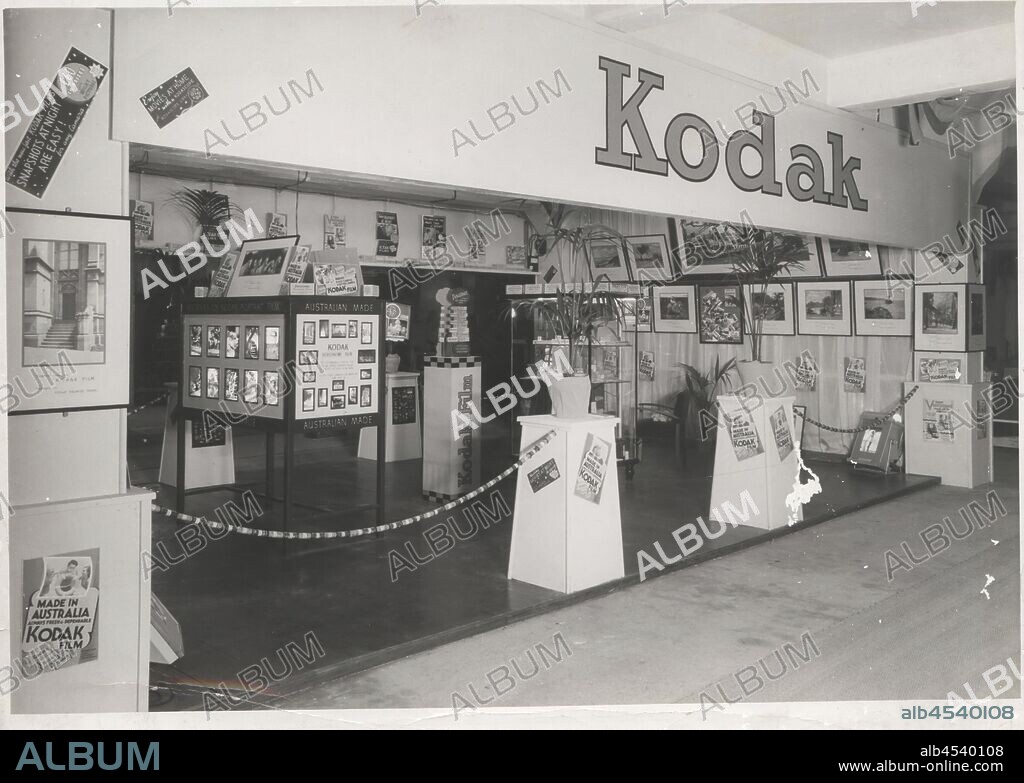 Photograph, Exhibition Stand, 'Australian Made', Chamber of Manufactures Exhibition, Geelong, 1938, Black and white photograph of a Kodak Australasia Pty Ltd 'Australian Made' exhibition stand / trade show booth at the Chamber of Manufactures Exhibition held in 1938 as part of the Geelong Centenary. There are freestanding glass cabinets, display boards, framed photographs and freestanding cardboard signs. The display is decorated with potted palms and an entranceway is created with plinths and a.