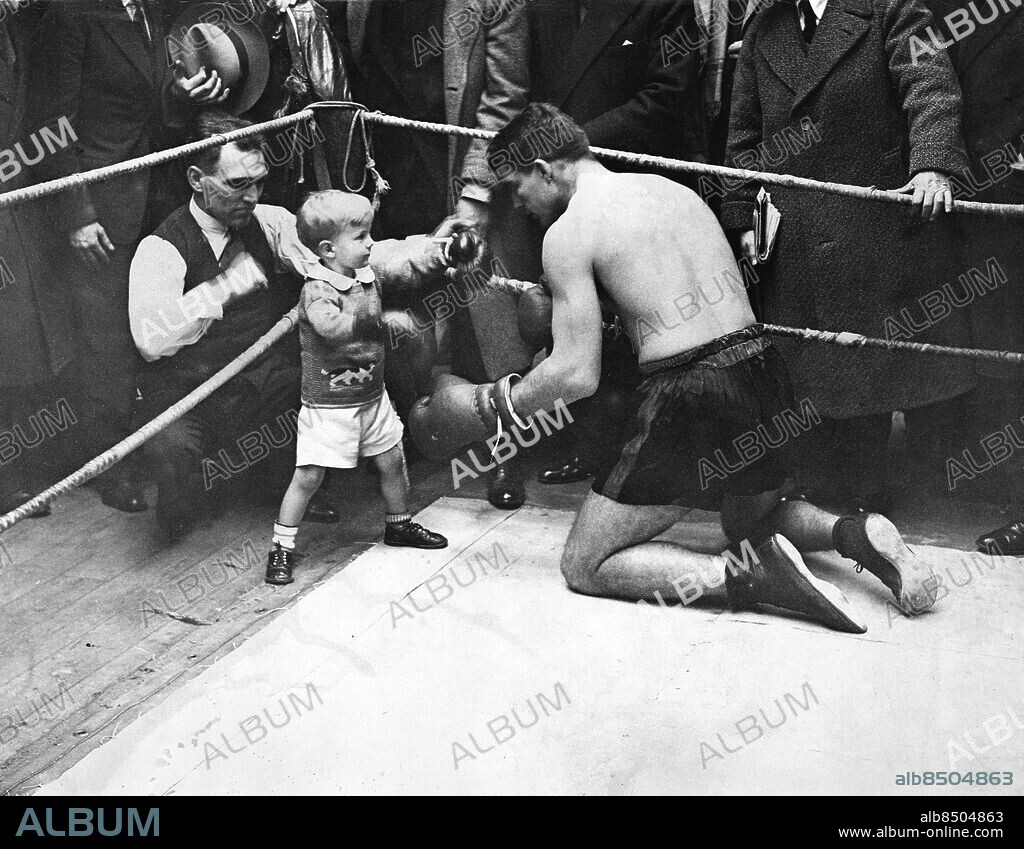 Orig. bildtext... YOUNG STRIBLING IN LONDON. 14.11.29. Young Stribling, the American boxer, who is to fight Primo Carnera, the giant Italian boxer, at the Albert Hall next week, is now instrict training in London. O.P.S. Young Stribling sparring with his little son in the ring, during practise this afternoon. Anm. Young Stribling, 1904-1933, amerikansk professionell tungviktsboxare. Sparrpartner Boxningsring Bar överkropp Boxningshandskar Sparring CD652 persons: YOUNG STRIBLING sites: LONDON;STORBRITANNIEN;USA*.