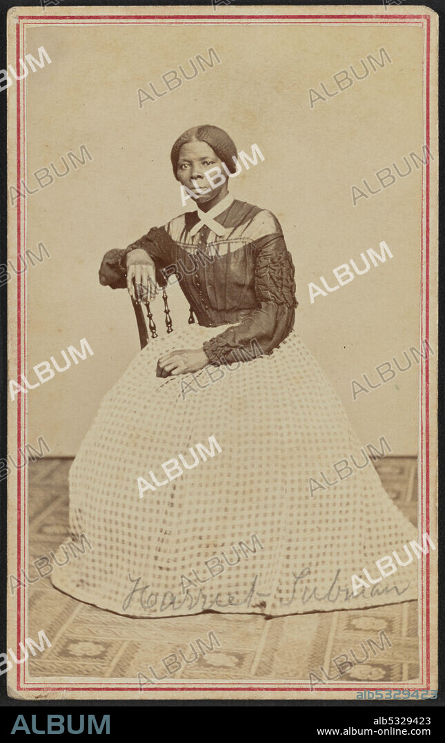 BENJAMIN F. POWELSON. African-American anti-slavery activist Harriet Tubman (c1820-1913) was born into slavery in Maryland. She escaped in 1849, became a leading Abolitionist and was active as a 'conductor' in the Underground Railroad, the network which helped escaped slaves to reach safety. She is positioned slightly turned to the right and gazes off camera. Her right hand rests on the back of a wooden chair and her left hand rests in her lap. A patterned carpet covers the floor and the wall or drop behind her is a blank light color. Tubman wears a black bodice that buttons at the center front and has dropped sleeves with heavy ruching and ruffled details on the sleeves. There is a panel of lighter fabric around the yoke, with the upper neck the same dark color as the body of the bodice. A white lace collar with short tails is crossed and pinned at the front of her neck. Her hair is parted at the center and gathered at the nape of her neck. Tubman's full skirt is made from a light and dark patterned gingham check. It drapes slightly on the floor and hides her feet completely. The text "Harriet Tubman" is handwritten in graphite across the bottom of her skirt. The print is mounted on card stock and a double-lined red border is printed around the front edges of the card framing the print.