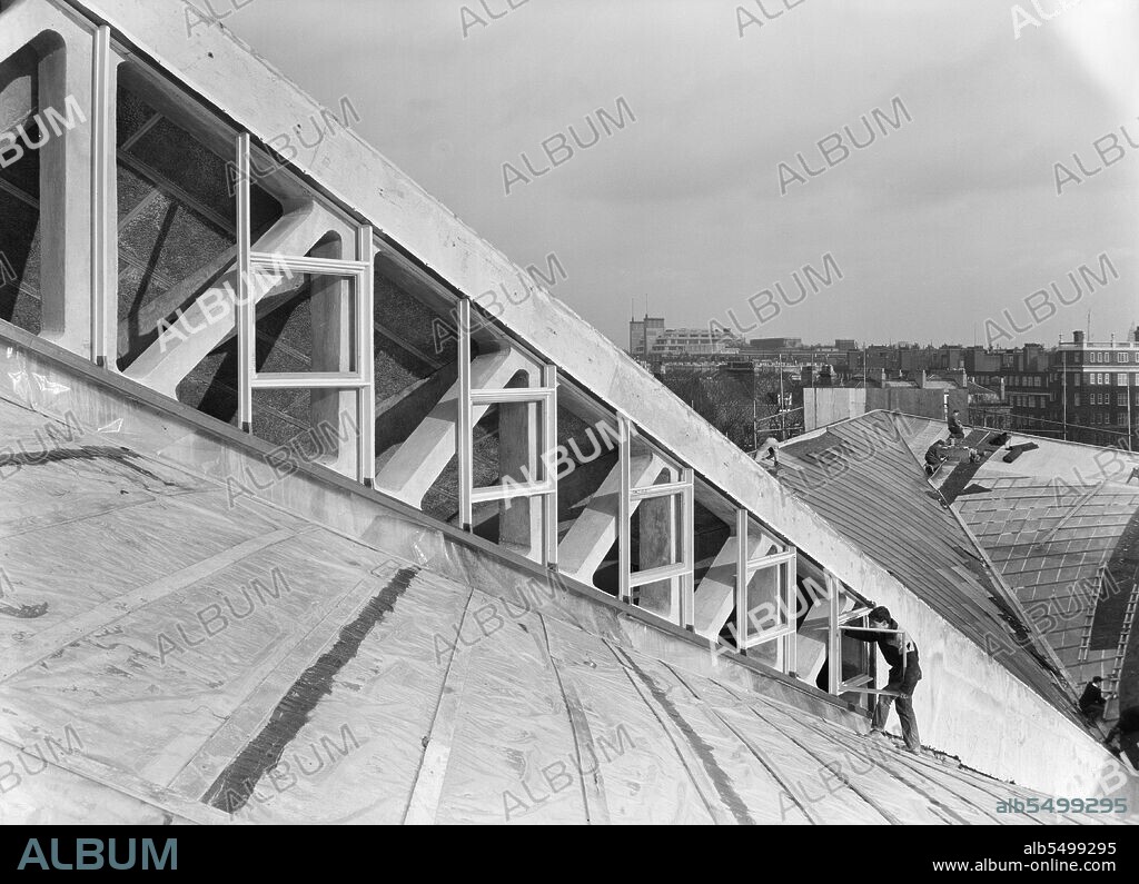 JOHN LAING PLC. Commonwealth Institute, Kensington High Street, Kensington, London, 06/04/1962. A worker installing window frames in the clerestory between the central and outer roof sections of the exhibition hall at the Commonwealth Institute. Laing built the Commonwealth Institute between October 1960 and October 1962 to replace the former Imperial Institute that was to be demolished to make way for new facilities at Imperial College.  The building  consisted of a four-storey administrative block housing a library, restaurant, board room and conference hall and a separate two-storey block containing a cinema with an art gallery above, but the focus of the project was the exhibition hall with its hyperbolic paraboloid roof, the first of its kind constructed in Great Britain.  The exhibition, designed by James Gardner, provided spaces where each of the Commonwealth nations could showcase their achievements and characteristics, primarily to school children as teaching aids to enliven history and geography lessons. The shell arch of the central roof section was of reinforced concrete, cast in situ using timber formwork with rough sawn boards to provide a textured internal surface whilst the four outer "warps" were constructed using precast beams and wood wool slabs, blocks of shredded timber bound together in a cement paste and left visible from the interior.  The entire roof was then clad in copper sheeting over a layer of vermiculite.  It covers an area of 33,700sqft, 183 feet square with the central section 93 feet square and ranges between 30ft high at its lowest and 80ft at the peaks.