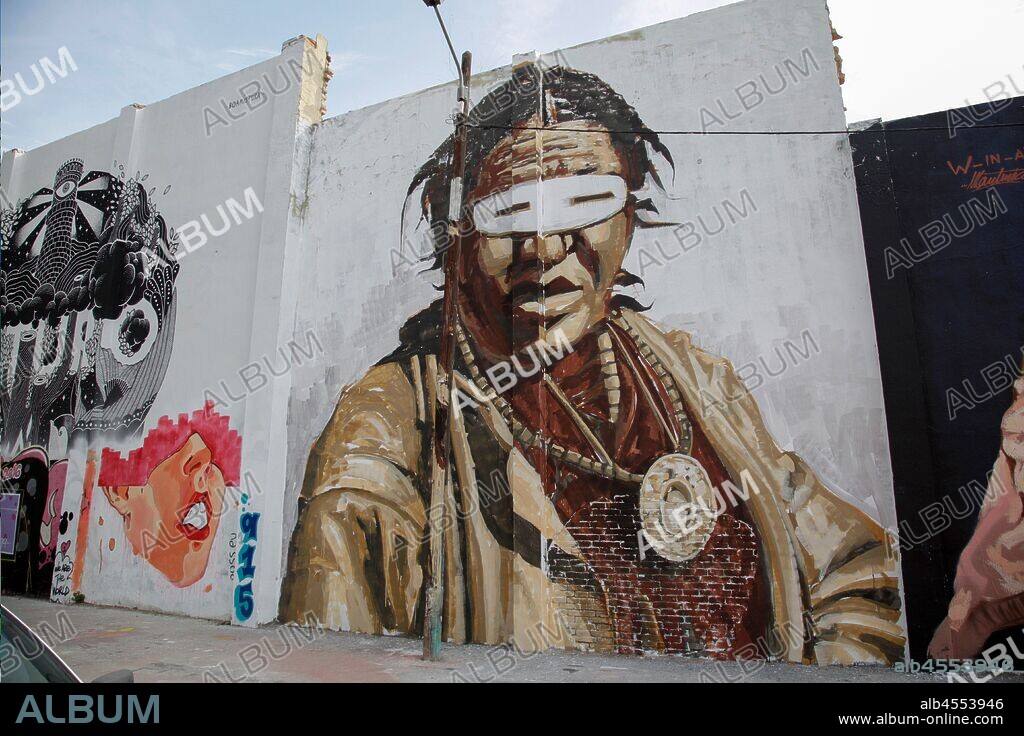 Pintura de Indio americano con chaqueta, antifaz y un gran colgante redondo, en tonos marrones, graffiti mural, calle Agricultura, Poblenou, Distrito San Martin, Barcelona, en 2016.
