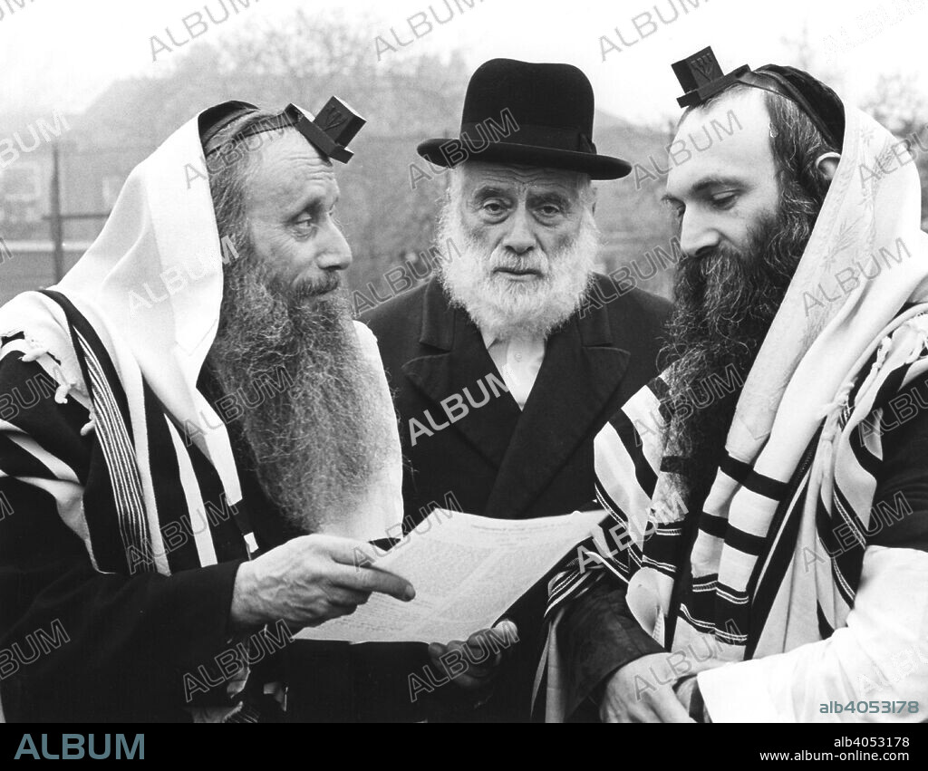 Hasidic Jews wearing tefillin and tzitzit, 1981. Tefillin are small boxes containing passages of religious text written on parchment scrolls. They are attached with leather straps, and worn during prayer. Tzitzit are woven shawls.