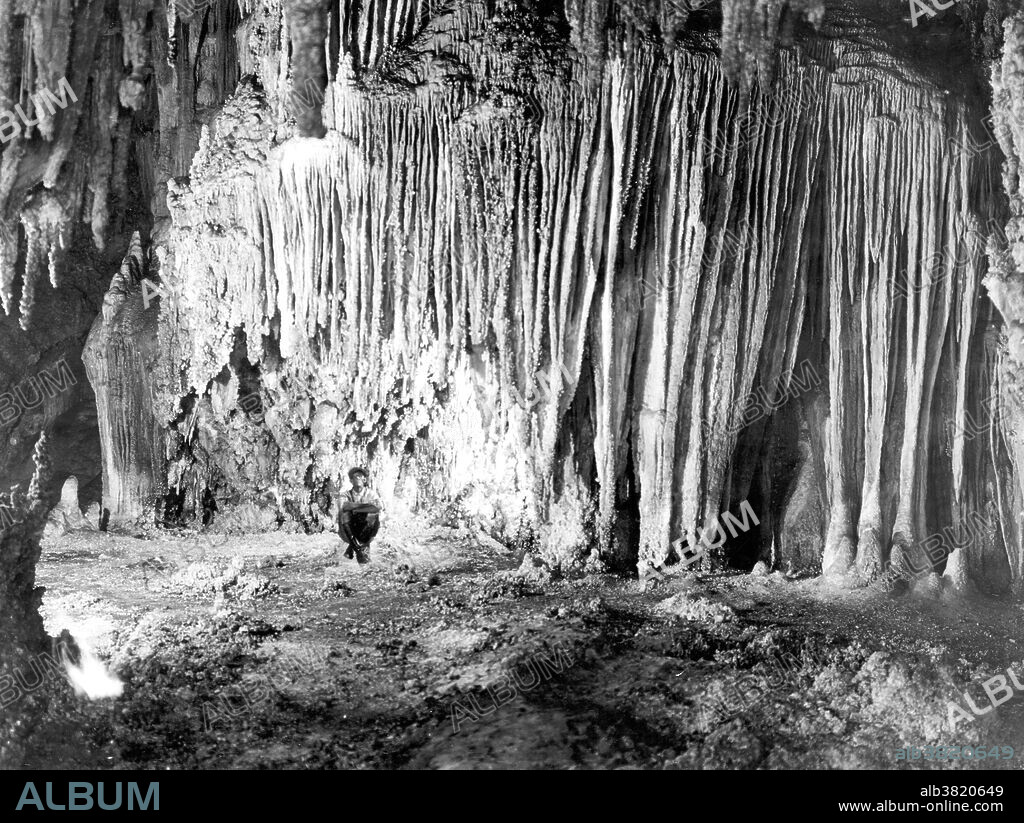 Photo was taken in 1923 inside Carlsbad Caverns National Park, New Mexico shows a wall in the chambers known as Shinav's Wigwam (from Shinav, a mythical deity of the Navajo Indians). The stalactites have grown together laterally to form a solid curtain-like mass. Carlsbad Caverns National Park is located in the Guadalupe Mountains in southeastern New Mexico. The primary attraction of the park is the show cave, Carlsbad Cavern. The Carlsbad Caverns are an extensive cave system with over 80 known caves. It originated 250 million years ago as erosion started on a reef in an (evaporated) inland sea. Visitors to the cave can hike in on their own via the natural entrance or take an elevator from the visitor center. Show caves - also called tourist caves, public caves, and in the United States, commercial caves - are caves that are managed by a government or commercial organization and made accessible to the general public, usually for an entrance fee. Unlike wild caves, they typically possess such features as constructed trails, guided tours, lighting, and regular opening hours.