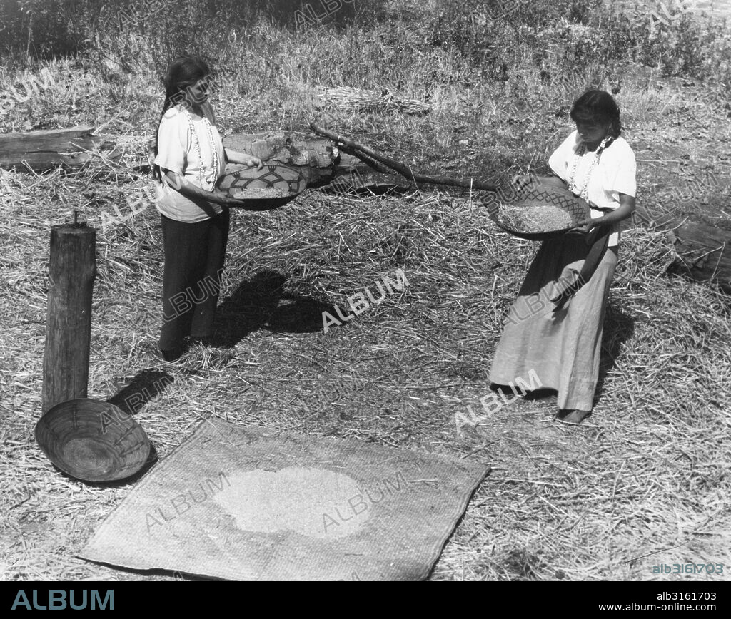 California: c. 1930 Two California Native American women winnowing wheat to separate the kernels from the chaff.