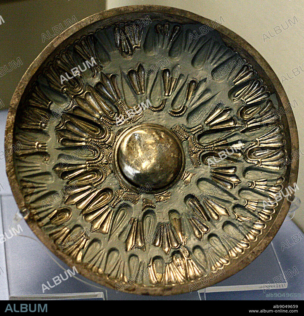 Sheet gold pendant showing a Cretan nature god, The figure wears a Minoan kilt, a tall head-dress, bracelets and earrings. He stands in the attitude of the 'Master of Animales'. In which a central figure flanked by animals demonstrates control over them. The curved, ridged elements surrounding the birds derive from stylised bulls' horns. Egyptian influence is seen in the lotus flowers among which he stands.