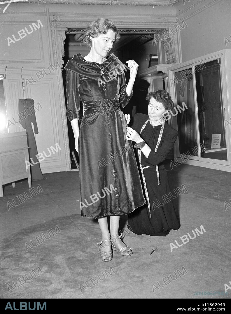 OPUS 1949. Lady Beecham ( Betty Humby , the pianist ) will show the Americans what Britain can do as one of the visual arts-dress designers -when she accompanies her husband ,Sir Thomas Beecham , the conductor , on his forthcoming visit to the United States . She has bought an extensive all-Britain wardrobe for the trip and will be ambassadress of fashion . Aware of the average American woman's love of shopping in stores , Lady Beecham has bought all her outfits at leading London stores . PICTURES SHOWS:- LADY BEECHAM having a fitting of a burgundy velvet dinner gown for her American trip. The gown has a swathed collar, framing the neck, and grouped velvet covered buttons down the front. Notes to fashion editors: This dress is by Marshall and Snelgrove. 17 October 1949.