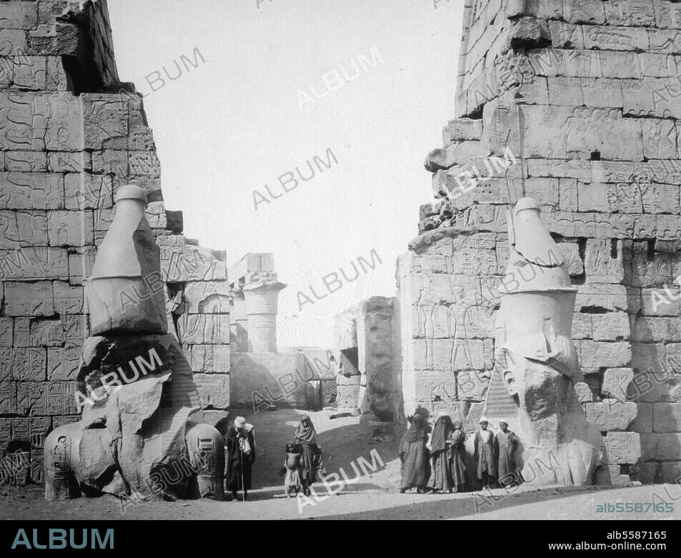 Luxor, Luksor, arab. El-Uksur (Oberägypten); Amun-Tempel (unter Amenophis III. c. 1400 v. Chr. erbaut; erweitert unter Ramses II.) / - Teilansicht: Erster Pylon mit Kolossalstatuen (Sitzstatue Ramses II., 19. Dynastie, 1290-1224 v. Chr.). vor der Freilegung. Photographie, anonym, um 1870. Paris, Privatsammlung.