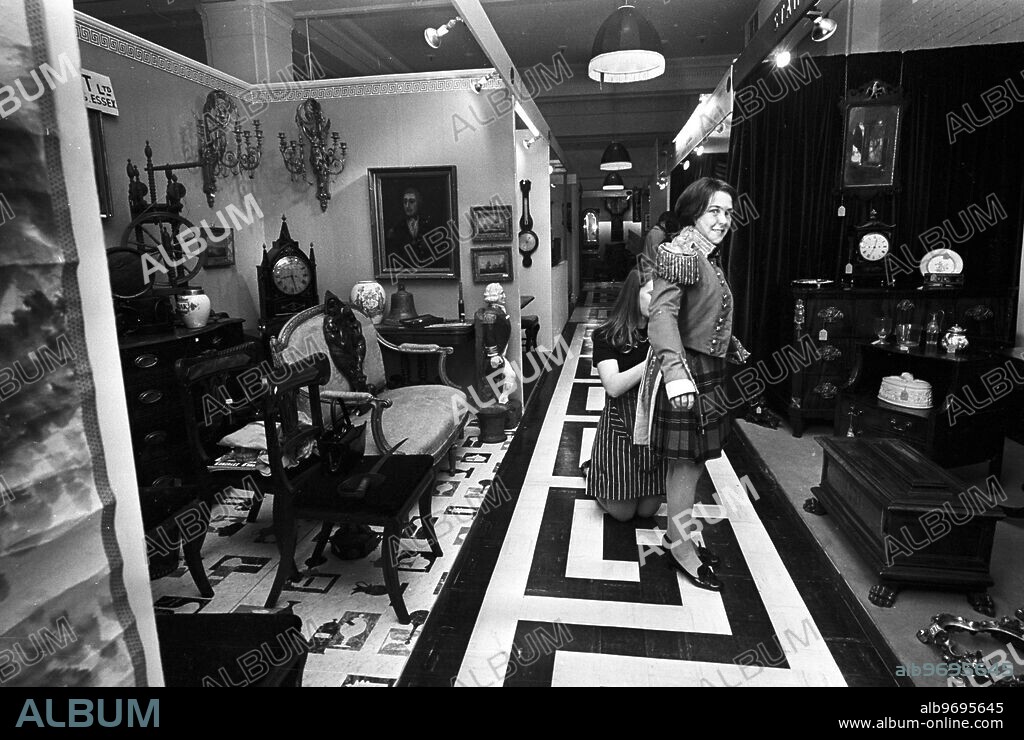 Inside the Antique Hypermarket, Kensington High Street, London, England, which houses nearly 100 shops and stalls. In the space bewteen the shops and stalls, two young girl workers, carry out some repairs on an old military uniform. Helpfullness is in the code of the Antique Hypermarket dealers. 2 December 1967.