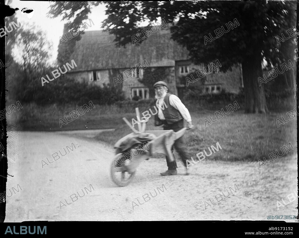 KATHERINE JEAN MACFEE. Moat Farmhouse, Whiston, Cogenhoe and Whiston, South Northamptonshire, Northamptonshire, 1920. A bearded elderly man pushing a wooden wheelbarrow across the road in Whiston, with Moat Farmhouse in the background.