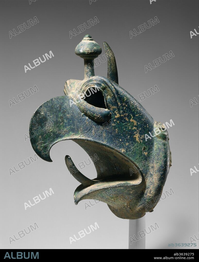Bronze head of a griffin. Culture: Greek. Dimensions: H. 10 3/16 in. (25.8 cm). Date: third quarter of the 7th century B.C..
From Olympia
Bronze cauldrons set on tripods or conical stands were among the most spectacular votive gifts dedicated in Greek sanctuaries from the eighth to the sixth centuries B.C. Cast-bronze griffins' heads often decorated the cauldron rims; they projected outward from the shoulder of the vessel on long necks made of hammered or cast bronze.  Some of the dedicated cauldrons were colossal. The Greek historian Herodotus describes one made for King Kroisos of Lydia that could hold 2,700 gallons and another dedicated on the island of Samos that was supported by huge kneeling figures. Over six hundred bronze griffins' heads from cauldrons are known today; most have been found at the sanctuary of Zeus at Olympia or at that of Hera on Samos. This enormous head is one of the finest.