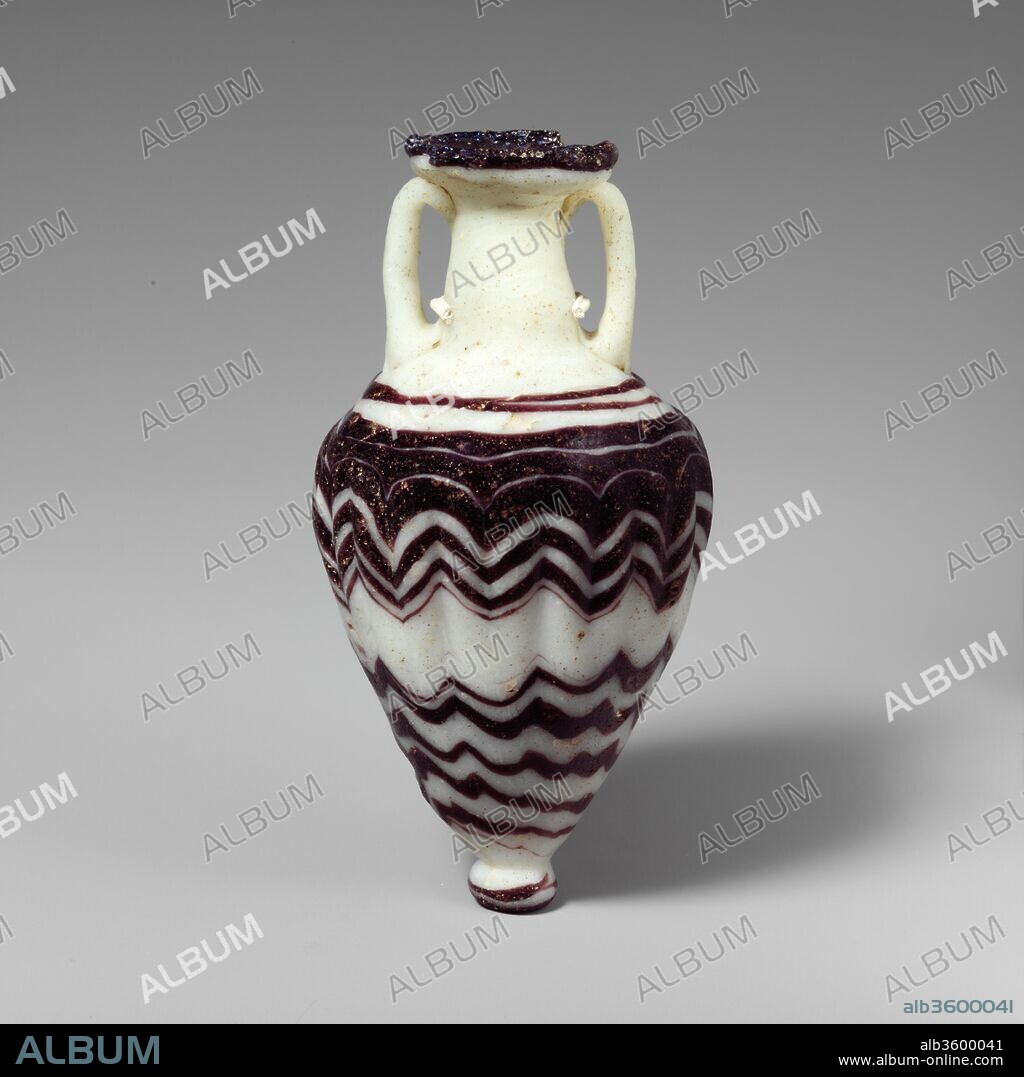 Glass amphoriskos (perfume bottle). Culture: Greek, Eastern Mediterranean. Dimensions: 4 3/4 × 2 7/16 in. (12 × 6.2 cm)
Diam. of rim: 1 5/16 in. (3.3 cm)
Diam. of base: 9/16 in. (1.5 cm). Date: late 6th-5th century B.C..
Opaque white, with handles and knob-base in same color; trails in translucent purple.
Broad, inward-sloping oval rim-disk with radiating tool marks on upper surface and small mouth; tall cylindrical neck tapering upward; broad, sloping shoulder; elongated ovoid body tapering to a point; slanting circular base-knob with rounded edge and flat bottom; two vertical strap handles applied at top of shoulder, drawn up vertically, turned in, and pressed on to underside of rim-disk and top of neck.
One uneven trail attached at edge of rim-disk; another trail wound round in an uneven spiral on shoulder, then tooled into a zigzag pattern on upper body, with twenty-two deep vertical tooling indents; a third trail applied to lower body, also tooled into an irregular zigzag pattern; finally, a fourth trail applied around edge and underside of knob-base.
Complete, but part of rim-disk broken and repaired, leaving one large chip and smaller hole; internal cracks in body; dulling, pitting, and faint iridescence.