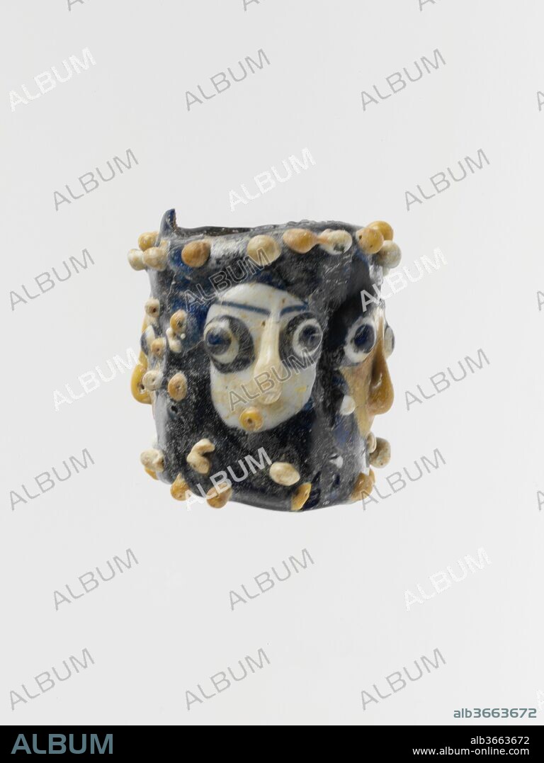 Glass face bead. Culture: Phoenician or Carthaginian. Dimensions: Diam.: 1 9/16 in. (4 cm). Date: late 5th-mid-2nd century B.C..
Translucent dark cobalt blue, with additions in opaque white and yellow and translucent blue.
Cylindrical, with rounded edge at both ends; large hole running vertically through bead.
Four faces, alternating with two in yellow and two in white, each made from a large added blob, augmented with applied nose, stratified eyes, eyebrows, ears, pendant earrings, and small round mouth, flanked above and below by irregular rows of knobs in white and yellow.
Broken and repaired, with some areas of fill, and a few knobs missing or damaged; some pitting and iridescent weathering on exterior, encrustation on interior of hole.
This large face bead is closely related to both eye beads and head pendants. Examples of both groups, which have been found throughout the Mediterranean region, are also displayed here [Gallery 171, Case 31].