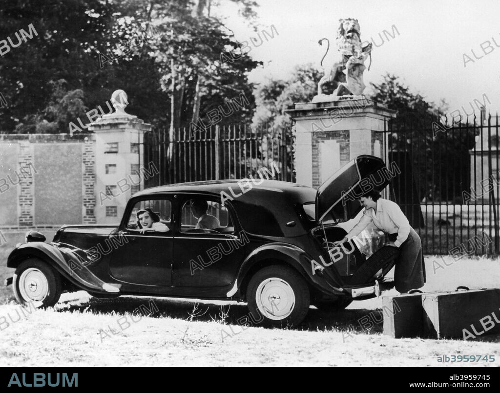1955 Citroën II Légère Berline car, (c1955?). A woman takes suitcases out of the boot. Behind are the entrance gates to a stately home, with a statue of a lion on the gatepost.