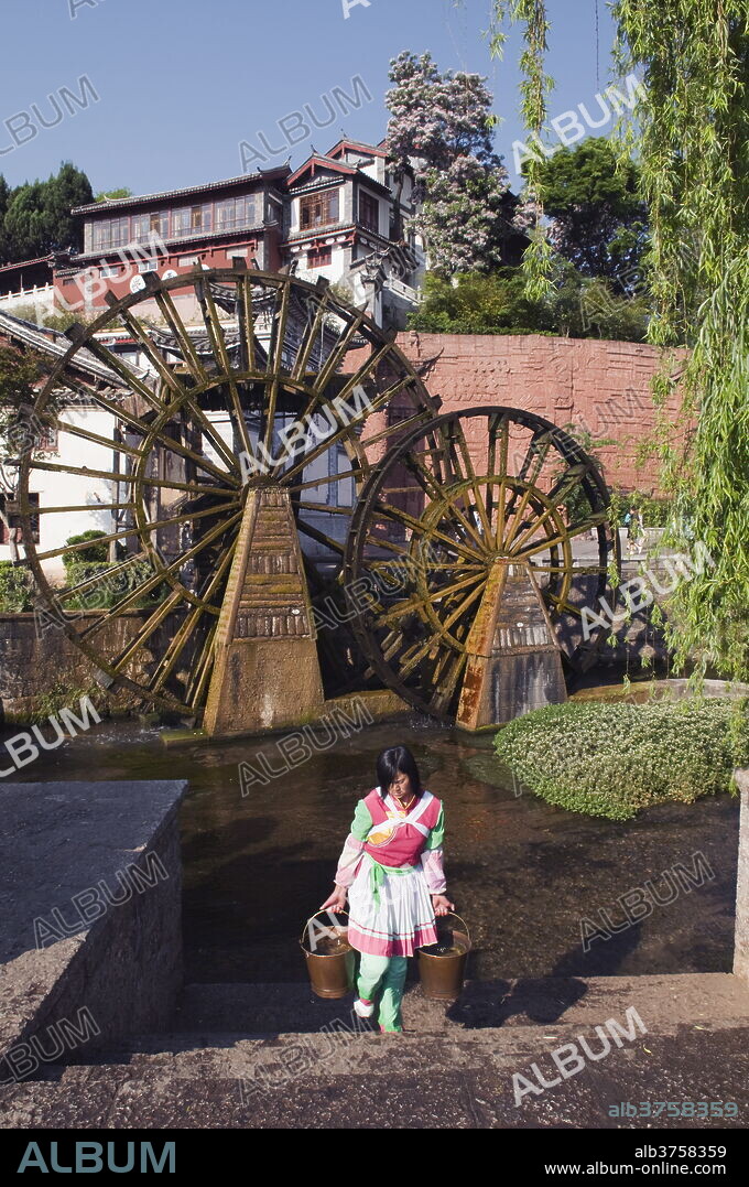 A Bai girl carrying buckets of water in front of a water wheel in Lijiang Old Town, UNESCO World Heritage Site, Yunnan Province, China, Asia.