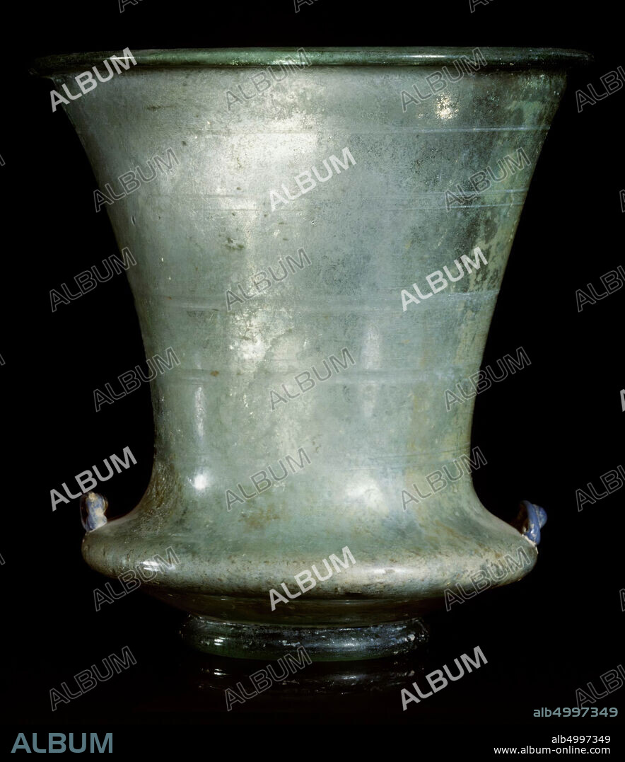 This perfectly shaped chalice is in the tradition of the showcup, which the Romans called carchesium. The light green glass is made of extremely thin-walled. The decoration consists of mini-ears of ultramarine glass on the shoulder and some ground horizontal bands on the upper part of the carchesium. The actual vase body consists of a layer and strongly convex part, which turns into an elegantly shaped, flared chalice. The shape is at the transition point of two worlds: on the one hand the piece stands in the tradition of the Roman showcup, on the other it points to the luster of Islamic glass art, with its mosque lamps in the same shape., Crockery, chalice, glass, H 14.6 cm, D 13.7 cm, Roman Period 400-700 ad, Turkey.