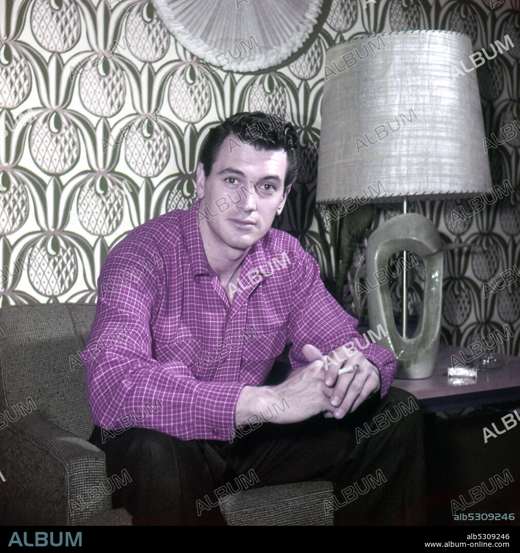 ROCK HUDSON. Circa 1955, Los Angeles, California, USA: American actor ROCK HUDSON sitting and smoking a cigarette while at home in Hollywood.