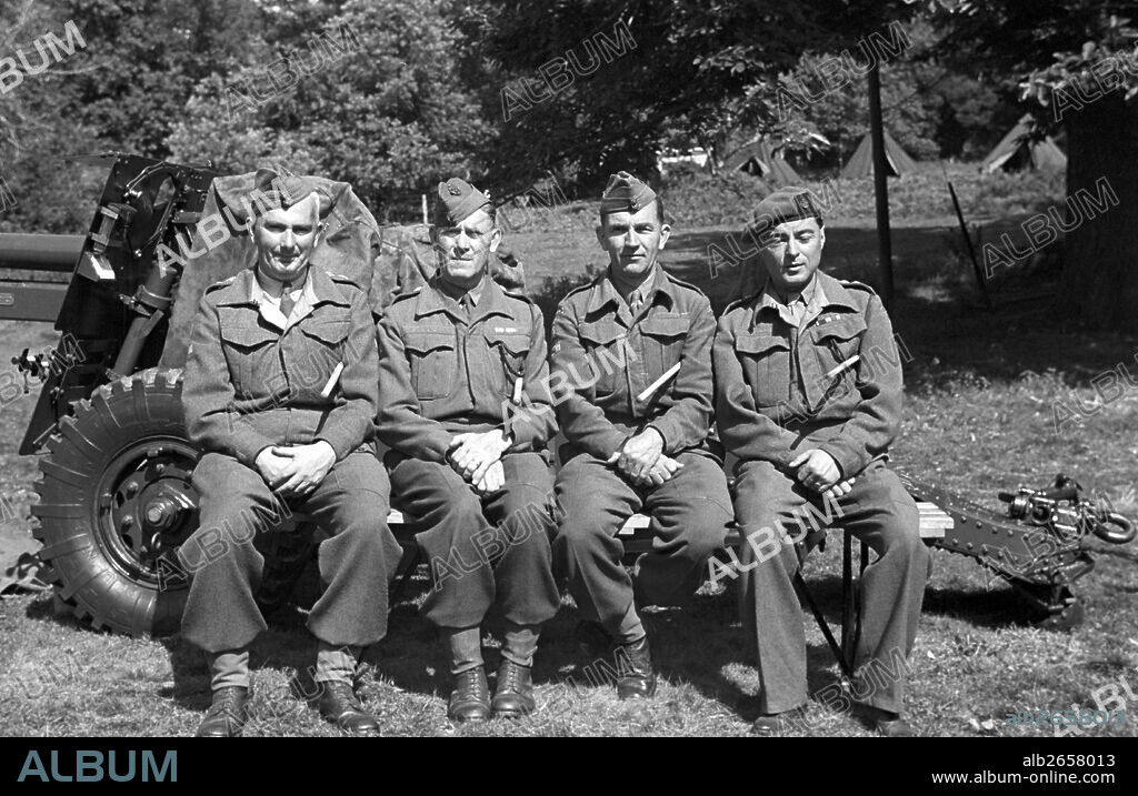 Four army officers seated on an artillery gun posing with swagger sticks. In the distance tents can be seen. Men in uniform, Soldiers.