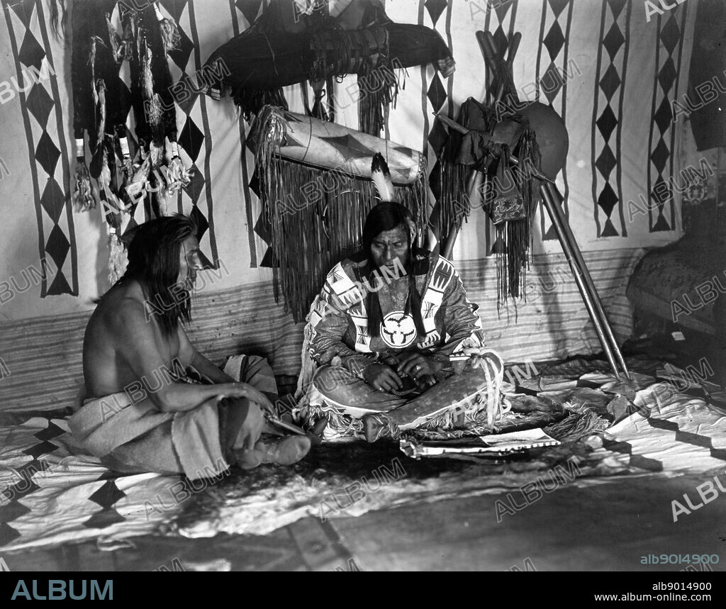 two Native American, Siksika (Blackfoot) men seated on blanket in a Medicine lodge in winter 1912.