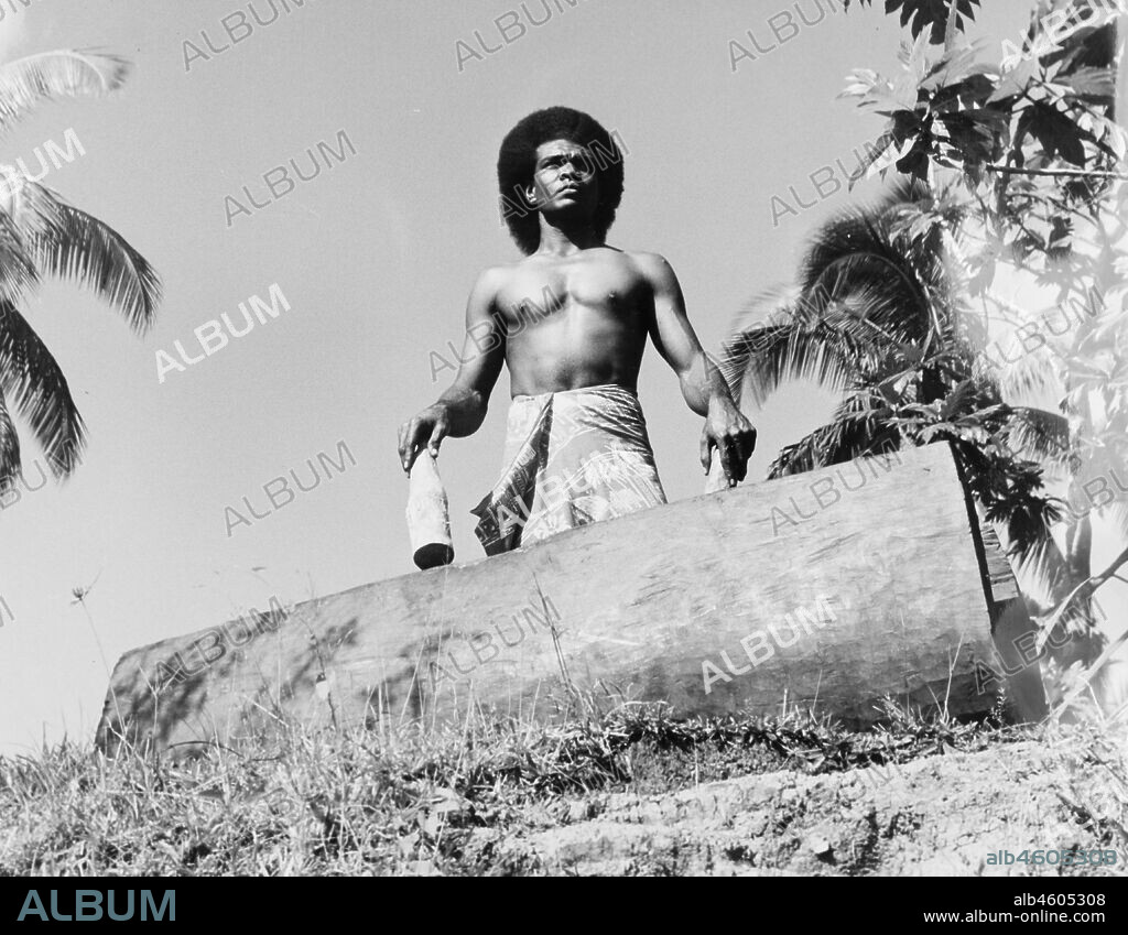 Fiji, A Fijian drummer with a 'lali'A Fijian man wearing a 'sulu' (wraparound skirt) stands over a traditional 'lali' (drum), holding a bottle-shaped wooden drumstick in each hand. Caption reads: The Fijian 'Lali' - a wooden drum made from a log hollowed like a trough - was used in ancient times to call warriors to a feast or to battle. When beaten with wooden mallets may be heard for many miles. Today it is used to call worshippers to church or elders to a village meeting, 1965. 2005/010/1/14/60.