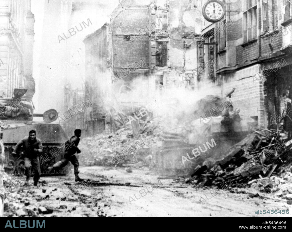 Dramatic Picture of the Battle For Cologne: During fierce fighting in the shadow of the Cologne Cathedral, a U.S. tankman, one leg blown off, lies on top of his was machine after he was blown from the turret by a German shell. While one First U.S. Army soldier (right) rushes to aid the wounded soldier, another runs for stretcher bearers. The wounded solider was the only members of the tank crew to survive the shell hit. The rubble-strewn street is the Komoodientrasse, running from Cathedral Squa.