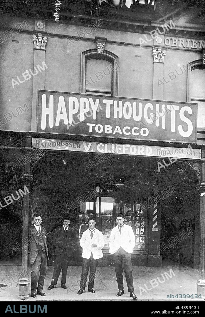 Negative - Port Melbourne, Victoria, circa 1920, Four men outside the shop of V. Clifford, hairdresser and tobacconist. A sign advertises 'Happy Thoughts Tobacco'. There is wrought iron lacework above the verandah of the shop.