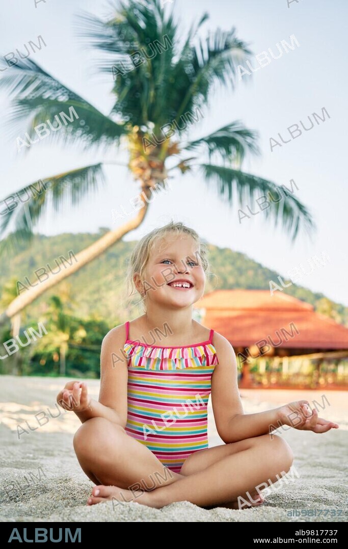 Pretty little girl meditating in Lotus Pose on sandy beach in summer vacation. Happy childhood concept.