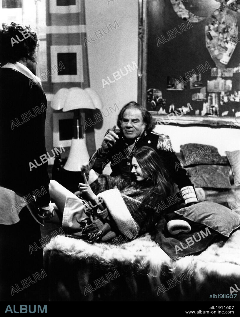 Lionel Stander and Sonia Petrovna seated on a sofa full of pillows. Lionel Stander, playing the role of count Eliogabalo and of Elia, Sonia Petrovna, playing the role of Alessandra Pizzullo, and the director Alfredo Giannetti (facing backwards) on the set of the movie 'Di mamma non ce n'è una sola'. Italy, 1974.