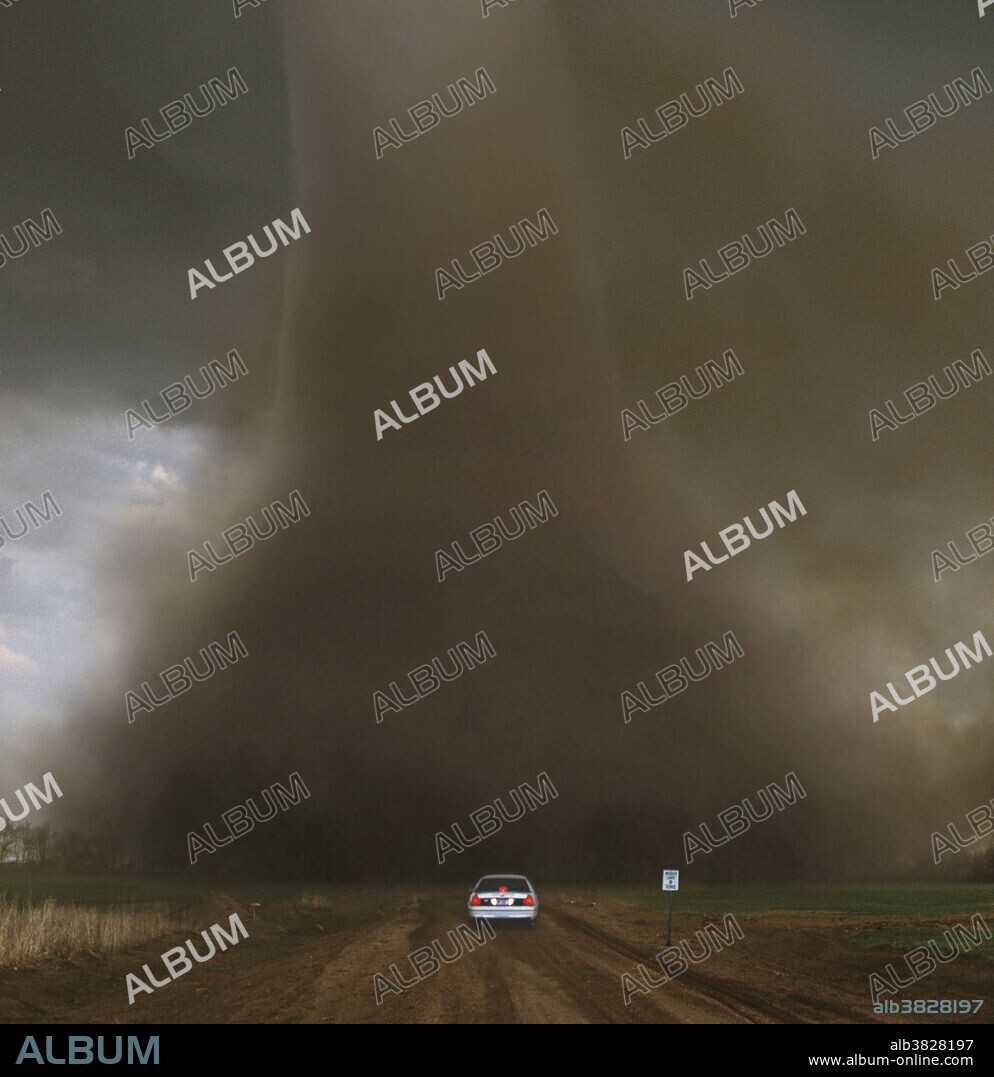 A tornado with large Liberty Bell-shaped debris cloud swirls across a dirt road less than 500 feet in front of a Kansas State Trooper. This image is a composite created from a high-resolution transparency (shot by Jim Reed) and low-resolution video (photographed by Katherine Bay). Except for overlapping the two medias, no manipulation has occurred. Distance, size, shape and color are authentic.