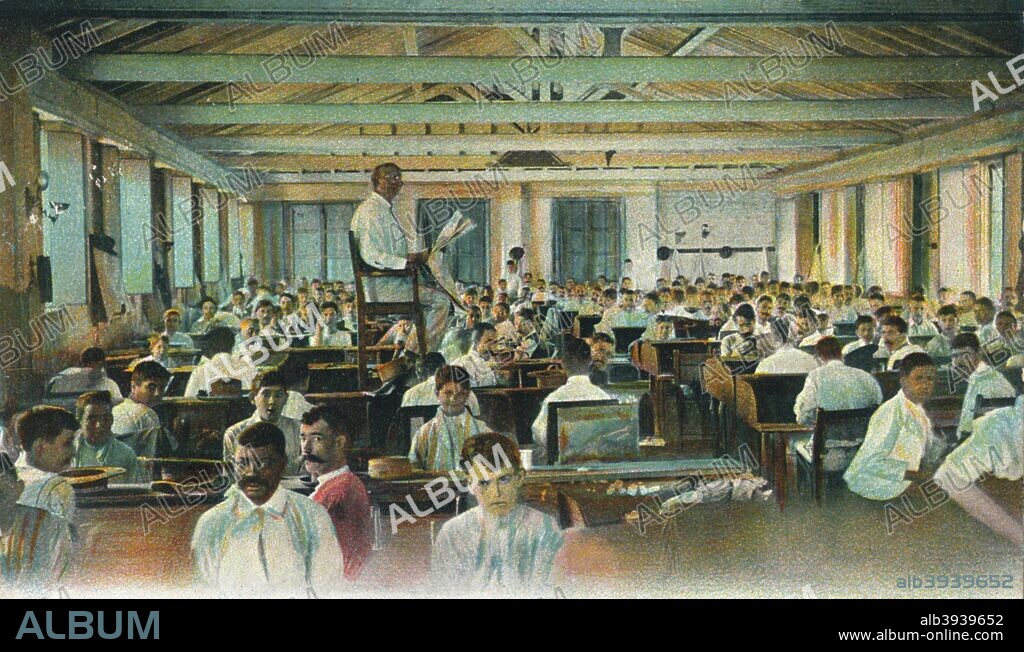 Lector reading to cigar rollers, Cigar Factory, Havana, Cuba. Readers are a traditional feature of Cuban cigar factories where the reader, often someone with radio experience, reads the news, stories or soap operas while the workers are busy rolling cigars. [The Rotograph Co., N. Y. City. (Germany.)].