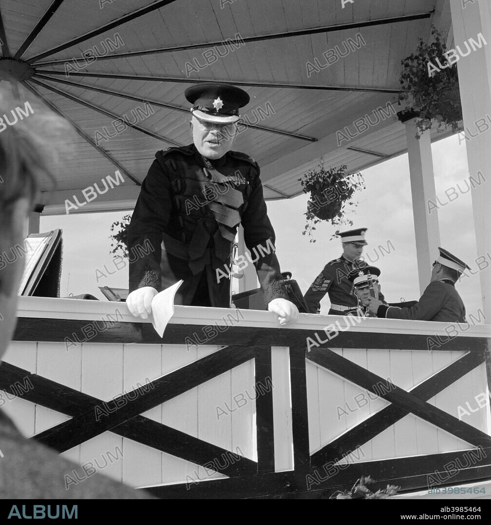 Conductor of the Band of the Coldstream Guards, Royal Agricultural Show, Newcastle upon Tyne, 1956 The conductor is leaning on the edge of the bandstand speaking to members of the public. Band members are seated behind.