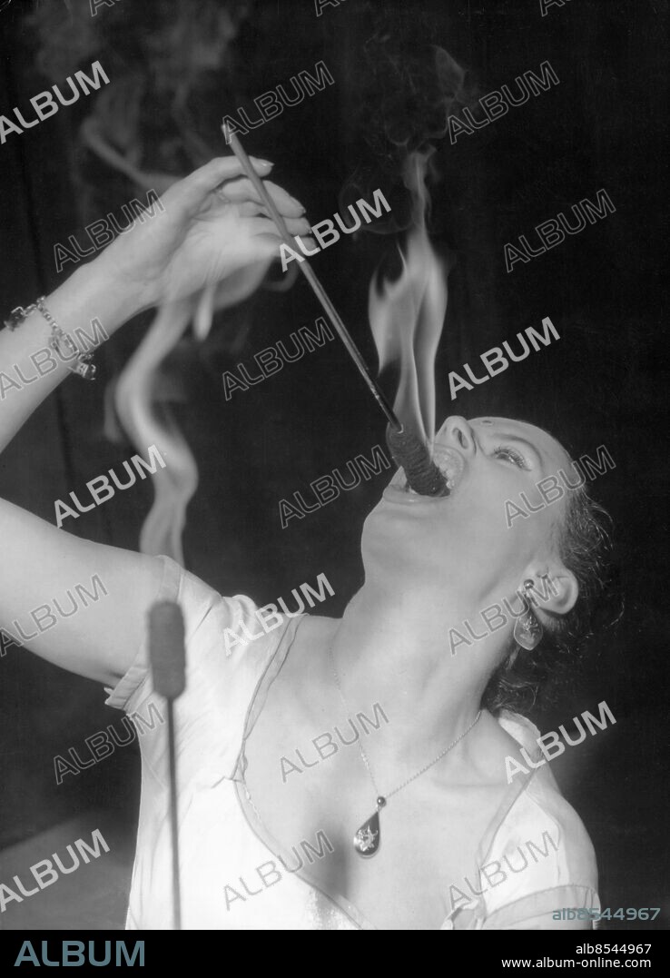 Orig. bildtext... SPITFIRE Lying on a bed of nails or walking bare foot through flames leaping up several feet around her, holds no terror for lovely 23 year-old British born Rahnee Motie, who claims to be the only white woman fakir in the world. I can do anything the fabulous fakirs of the East can do, Rahnee is reported to have said. Why should a beautiful girl want to do such things? She explains, her husband was the fire-eater of the family, but illness forced him to give it up, so she carries on in his place. She is now appearing in the 12th Annual Festival of Magic at the Scala Theatre, London. Photo shows: Rahnee demonstrates her fire-eating act. 14th. Oct. 1959. Anm. Fakirer Facklor Gapar Halsband Örhängen Lågor Farligt yrke CD526 persons: RAHNEE MOTIE sites: LONDON;STORBRITANNIEN.