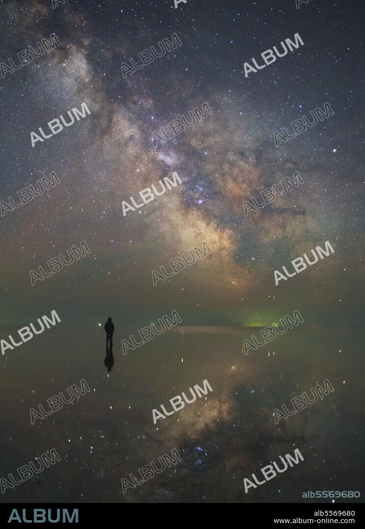 Man stands alone on Lake Elton in Russia under the center of the Milky Way and the stars reflected in the lake.