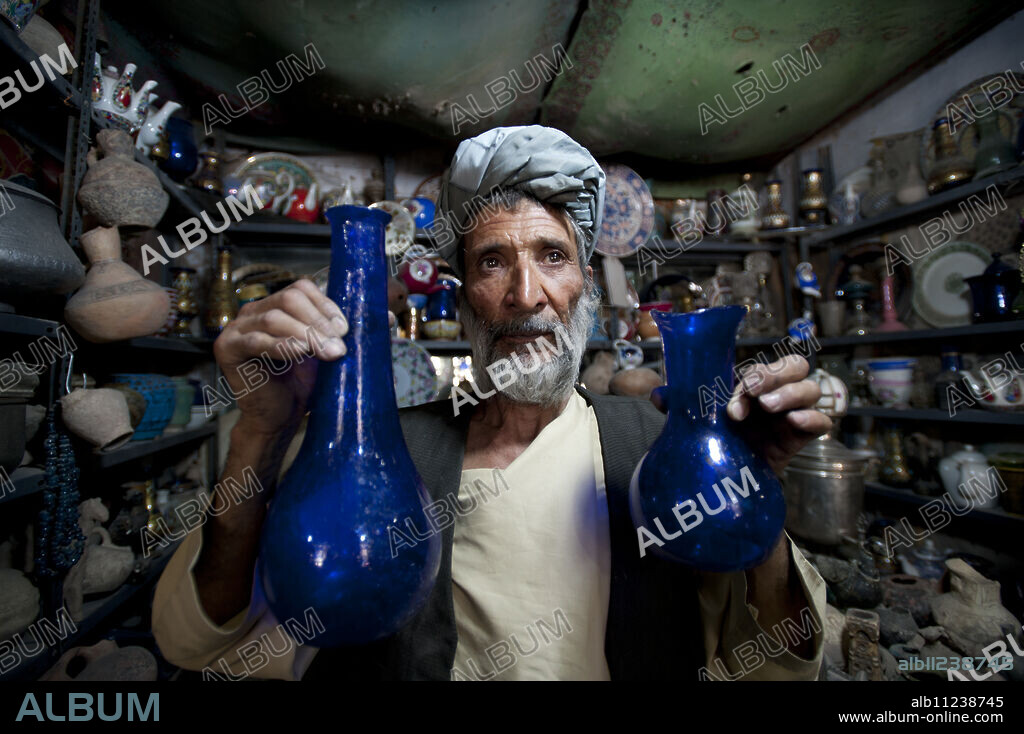 A glass blower holds up blue glass gourd-shaped vases in a trinket shop in Herat, Afghanistan, Asia.