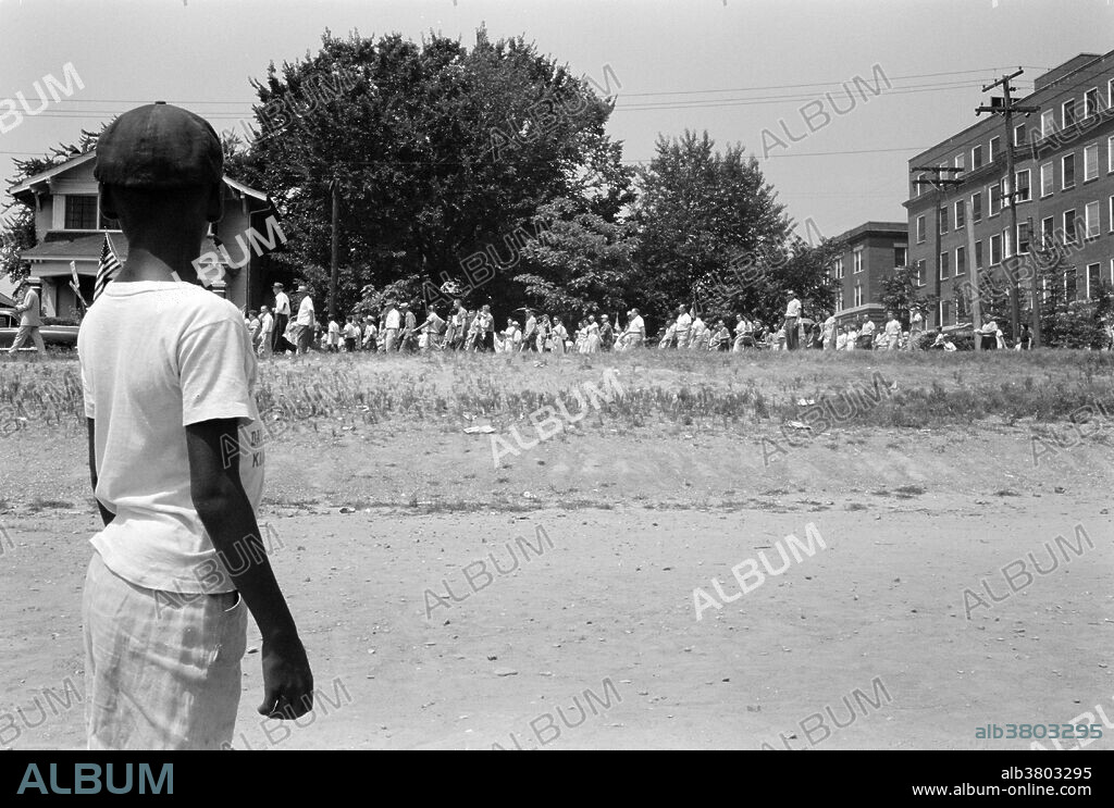 Entitled: "Little Rock, 1959. Mob marching from capitol to Central High" shows a young African-American boy watching a group of people, some carrying American flags, march past to protest the admission of the "Little Rock Nine" to Central High School. The Little Rock Nine were a group of nine African-American students enrolled in Little Rock Central High School in 1957. Their enrollment was followed by the Little Rock Crisis, in which the students were initially prevented from entering the racially segregated school by Orval Faubus, the Governor of Arkansas. After the 13th Amendment abolished slavery in America, racial discrimination became regulated by the so-called Jim Crow laws, which mandated strict segregation of the races. This legislation that mandated segregation lasted to the mid-1960s. Institutionalized racial segregation was ended as an official practice by the efforts of such civil rights activists as Clarence Mitchell, Rosa Parks, and Martin Luther King Jr., working during the period from the end of WWII through the passage of the Voting Rights Act and the Civil Rights Act of 1964. Photographed by John T. Bledsoe, August 20, 1959.