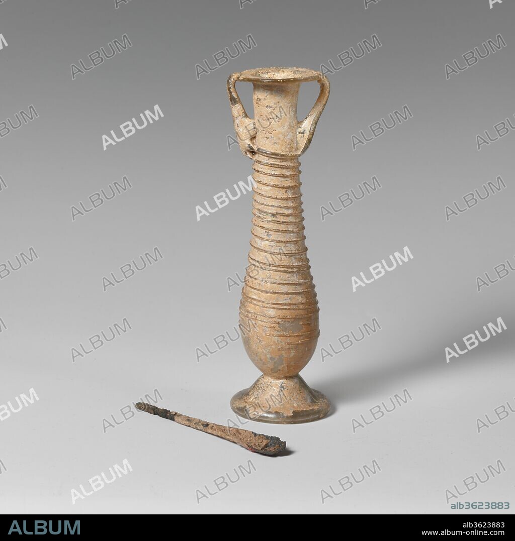 Glass cosmetic flask (kohl tube). Culture: Roman, Syrian. Dimensions: Overall: 5 9/16in. (14.1cm). Date: 4th century A.D..
Colorless with pale green tinge; foot, handles, and trail in same color.
Broad, horizontal rim folded out, over, and in; cylindrical neck, joining imperceptibly with slender, piriform body; applied conical foot, with folded tubular edge; deep kick in bottom and pontil mark; two rod handles applied in claw pads to neck, drawn up and out, and then turned in and pressed onto lip of rim.
Single fine trail applied as a pad on lower body, drawn up and wound in a spiral twenty-one times around body and neck, ending under one of the handles.
Complete, but crack in neck and upper body, and some parts of trail missing; some bubbles; dulling, slight pitting, thick creamy brown weathering, and limy encrustation.
Associated with the flask is a small bronze spatula, suitable for spooning the contents out of the deep flask.
